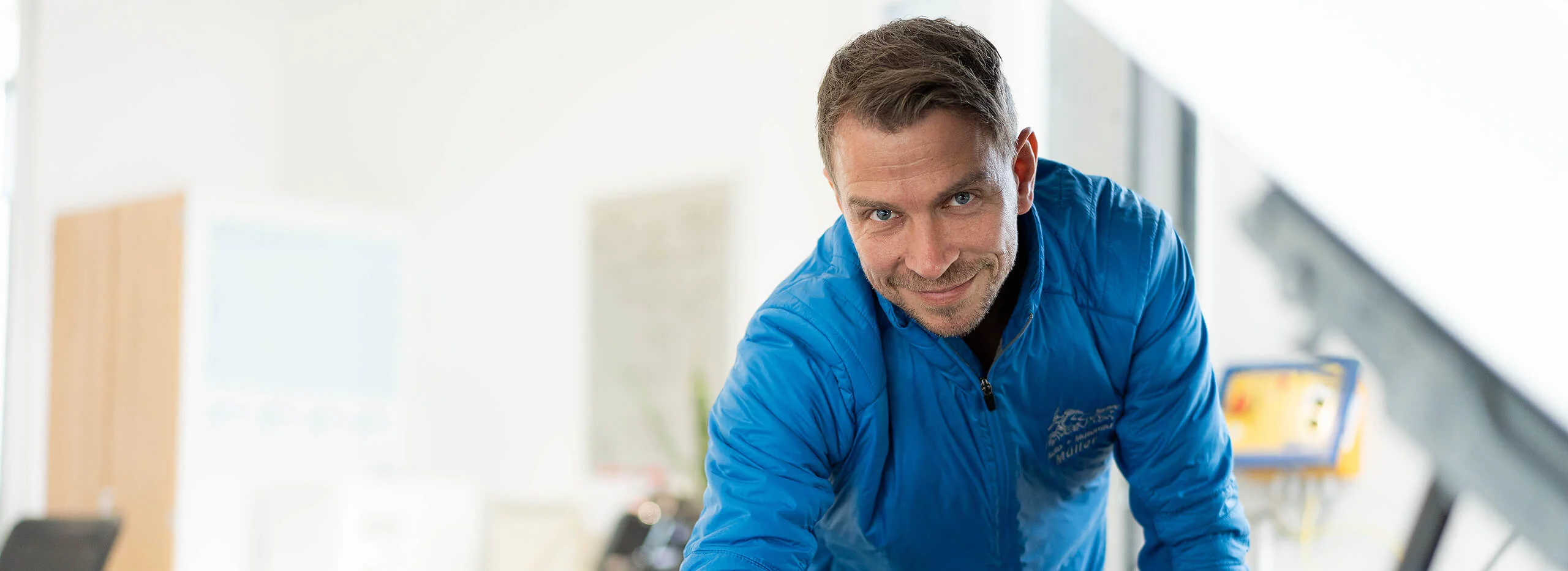 Ein lächelnder Mann in blauem Overall blickt in die Kamera, in einem hellen, unscharfen Innenraum.