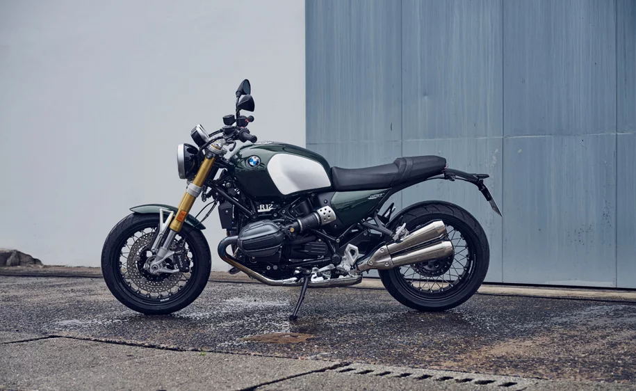  Dunkelgrünes BMW R 12 nineT Motorrad auf nassem Pflaster vor grauer Wand und blauer Tür.