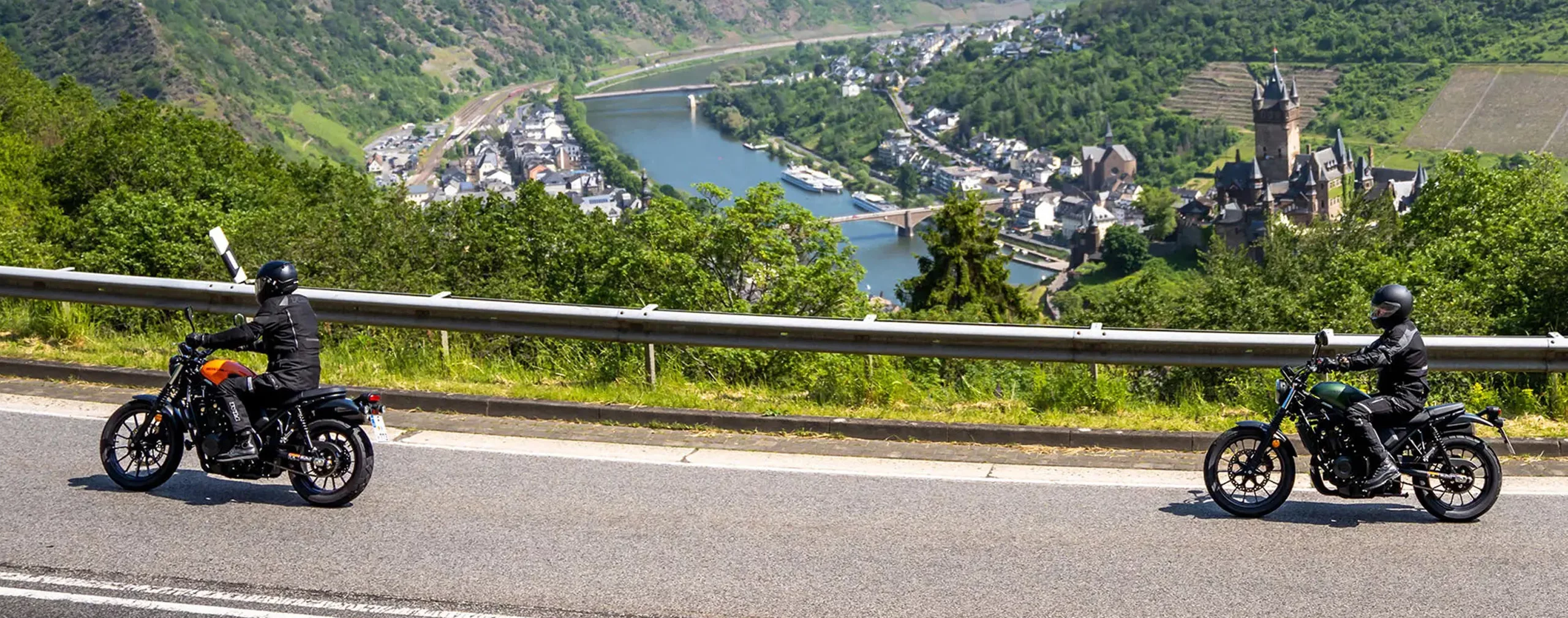 Zwei Motorradfahrer auf klassischen BMWs fahren auf einer Bergstraße mit Fluss- und Stadtblick.