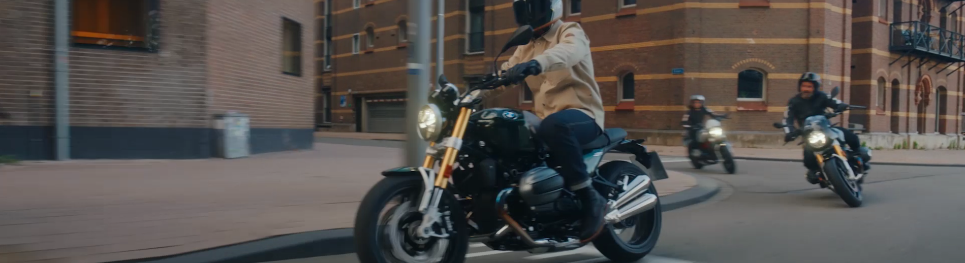 Ein Foto in einem breiten Panorama-Format, das eine Gruppe von drei Motorradfahrern zeigt, die in einer Stadt die Straße entlangfahren. Die Motorräder sind im Vintage-Stil gehalten. Im Vordergrund fährt ein Motorradfahrer in einer beigen Jacke auf einem schwarzen Motorrad mit goldfarbenen Elementen. Dahinter, auf der rechten Seite, fährt ein weiterer Fahrer mit Helm, der lächelt, auf einem Motorrad, das ebenfalls goldfarbene Elemente hat. Ein dritter Fahrer ist weiter hinten zu sehen. Die Szene spielt an einem sonnigen Tag in einer städtischen Umgebung mit Backsteingebäuden im Hintergrund.