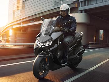 Das Foto zeigt den BMW C 400 GT in Fahrt auf einer mehrspurigen Stadtstraße während der Goldenen Stunde. Ein Fahrer mit weißem Helm steuert den Scooter an modernen Glasgebäuden und einer Brückenkonstruktion vorbei. Besonders markant ist hier die geschlossene, aerodynamische Frontpartie mit den leuchtenden LED-Tagfahrringen, die dem GT ein sehr präsentes und hochwertiges Gesicht verleihen. Die fließenden Linien und das hohe Windschild unterstreichen den Fokus auf Windschutz und Langstreckentauglichkeit.