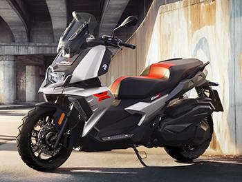 Das Foto setzt den BMW C 400 X in einer urbanen Szenerie unter einer Betonbrücke in Szene. Das Fahrzeug präsentiert sich hier in einer hellen Metallic-Lackierung (Zenith Blue oder Alpinweiß), die einen starken Kontrast zur zweifarbigen Sitzbank in Rot und Schwarz bildet. Besonders auffällig ist das rote „X“-Grafikelement an der Seitenverkleidung, das den sportlichen Charakter unterstreicht. Die Lichtstimmung ist warm und betont die dynamische Linienführung sowie die robusten Reifen, die für den Stadtverkehr optimiert sind.