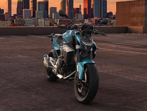  Auf dem Bild ist eine BMW F 900 R abgebildet, ein mittelgroßes, hellblaues und gelbes Motorrad, das auf einer Dachterrasse steht.  Das Motorrad ist in einem warmen, sonnigen Licht und steht seitlich zur Kamera. Es ist sauber und glänzt, als wäre es brandneu. Im Hintergrund befindet sich eine städtische Skyline bei Sonnenuntergang.