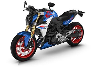 Das Motorrad steht in einer Dreiviertel-Ansicht von vorne rechts. Der aggressive Look wird durch den markanten LED-Scheinwerfer und die golden eloxierten Gabelbeine betont. Besonders auffällig sind die knallrot lackierten Gussräder vorne und hinten, die einen starken Kontrast zur blau-weiß-roten Verkleidung und dem schwarzen Motorblock bilden.