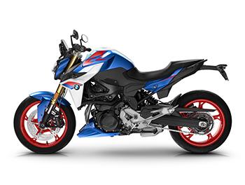 Das Bild zeigt die komplette linke Seite des Motorrads im M-Sport-Farbschema. Die gestreckte, dynamische Silhouette mit der ansteigenden Heckpartie und dem schlanken Heckträger ist gut zu erkennen. Die rote Felge hinten sticht hervor, ebenso wie die blaue Kühlerverkleidung mit dem BMW-Logo und der seitlich angebrachte Kennzeichenhalter. Der mächtige Vierzylinder-Motor ist schwarz gehalten.