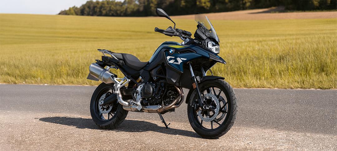  Auf dem Bild ist eine BMW F 800 GS abgebildet, ein mittelgroßes, dunkelblaues und gelbes Motorrad, das auf einer asphaltierten Straße steht.  Das Motorrad ist in einem hellen, sonnigen Licht und steht seitlich zur Kamera. Der Hintergrund ist eine grüne Wiese mit einigen gelben Blumen. Die Straße biegt leicht nach links ab und verläuft den Hang hinauf.