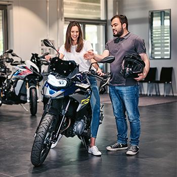 Das Bild zeigt eine freundliche Beratungssituation in einem modernen Motorrad-Showroom. Im Zentrum sitzt eine lächelnde Frau auf einer dunkelblauen Reiseenduro, während ein Verkäufer oder Berater neben ihr steht, einen Helm im Arm hält und ihr Details zum Fahrzeug erklärt. Im Hintergrund sind weitere Motorräder derselben Kategorie sowie eine helle, großzügige Werkstatt- oder Verkaufsatmosphäre zu erkennen. Die Szene wirkt einladend und professionell, wobei der Fokus auf der Vorfreude und der persönlichen Betreuung beim Motorradkauf liegt.