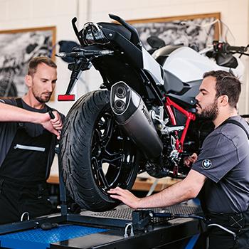 Dieses Motiv zeigt zwei Mechaniker bei der konzentrierten Arbeit in einer professionellen Werkstatt. Sie führen gerade einen Reifenwechsel an einer weiß-roten BMW Reiseenduro durch, die sicher auf einer Hebebühne aufgebockt ist. Die Szene vermittelt technisches Know-how und Präzision, während im Hintergrund Werkzeuge und Bilder an den Wänden für eine authentische Werkstatt-Atmosphäre sorgen.