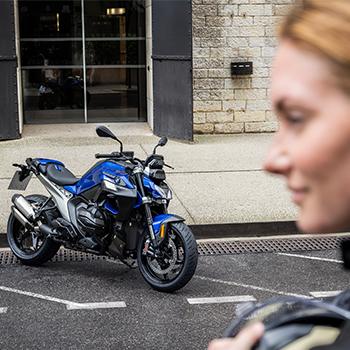 Das Bild fängt die Erwartung vor einer Probefahrt ein: Im linken Bildbereich steht eine markante, blau-schwarze BMW Roadster auf einer gepflasterten Fläche vor einem modernen Gebäude. Rechts im Vordergrund sieht man unscharf das Profil einer Frau in Motorradbekleidung, die den Blick auf die Maschine richtet. Die Komposition unterstreicht den Moment kurz bevor es auf die Straße geht, und setzt das sportliche Design des Bikes in den Fokus.