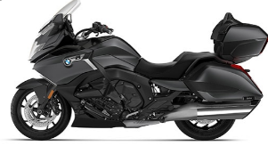 Ein schwarzes oder sehr dunkles Motorrad von BMW, das aussieht wie ein Tourenmodell, fotografiert schräg von der Seite vor einem weißen Hintergrund. Es hat ein großes Topcase und seitliche Koffer. Unter dem Bild steht der Modellname K 1600 Grand America in großer, fetter, schwarzer Schrift.