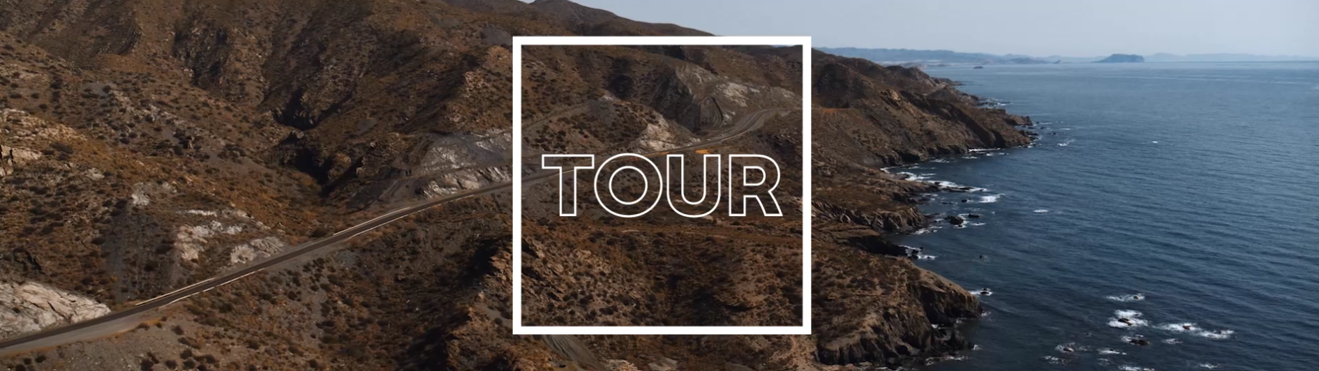 Motorradtour auf einer Küstenstraße entlang felsiger Berge mit Blick auf das Meer, Schriftzug ‚Tour‘ im Vordergrund – ideal für Roadtrip und Reisen.