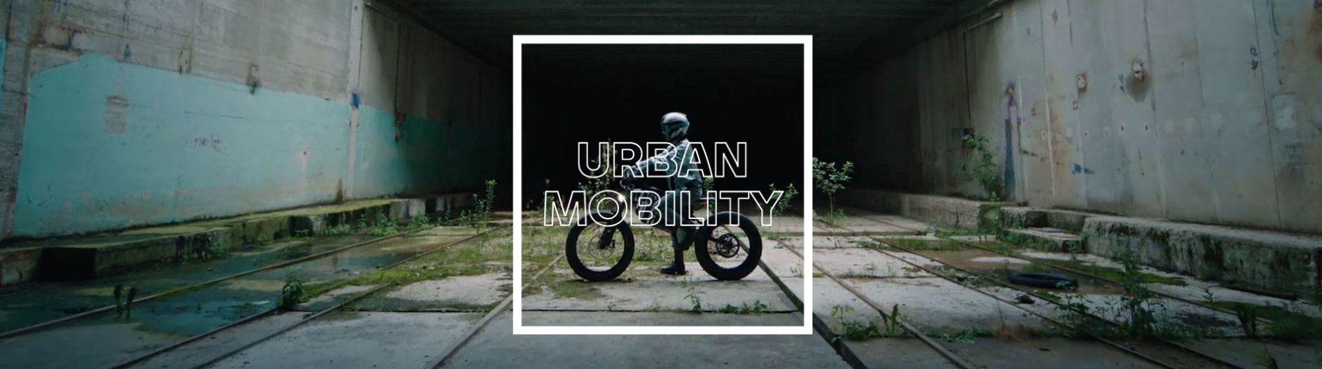  Ein Motorradfahrer in voller Montur sitzt auf einem futuristisch wirkenden Elektromotorrad. Sie stehen in einer verlassenen, urbanen Umgebung, die wie ein alter Tunnel oder ein verlassener Abwasserkanal aussieht. Die Wände sind aus Beton, teils mit Graffiti besprüht, und auf dem Boden wächst Unkraut. Die Szene ist dunkel und industriell. In der Mitte des Bildes befindet sich ein breiter, weißer Rahmen mit dem Text „URBAN MOBILITY“ in Großbuchstaben.
